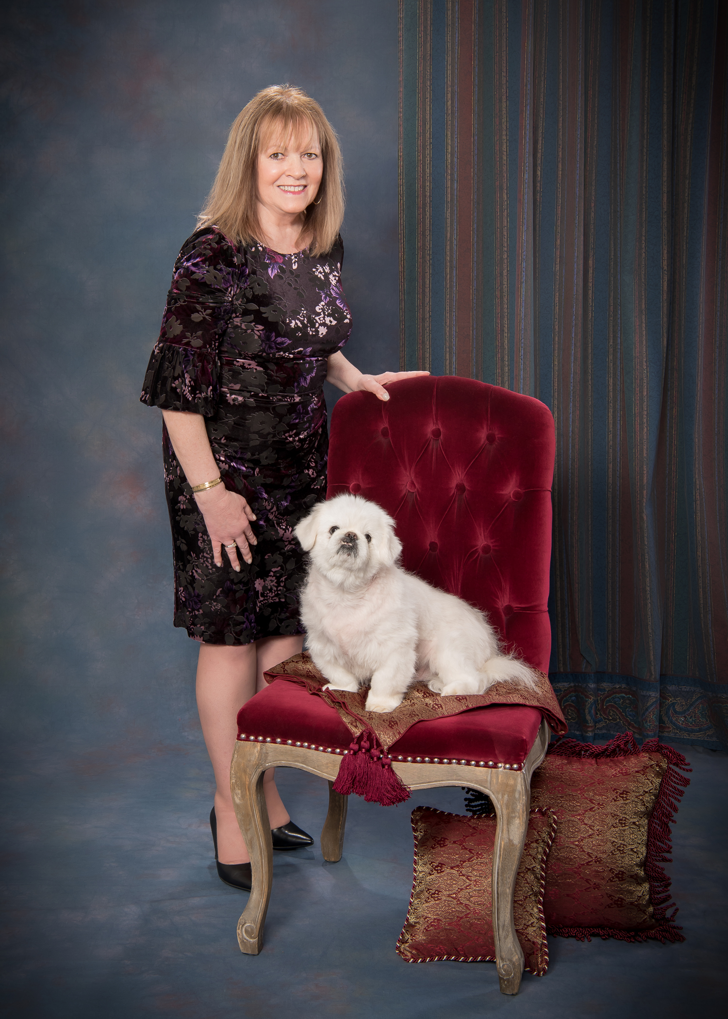 Picture of a standing woman next to her white dog seated on a red velvet chair in a South Jersey studio.