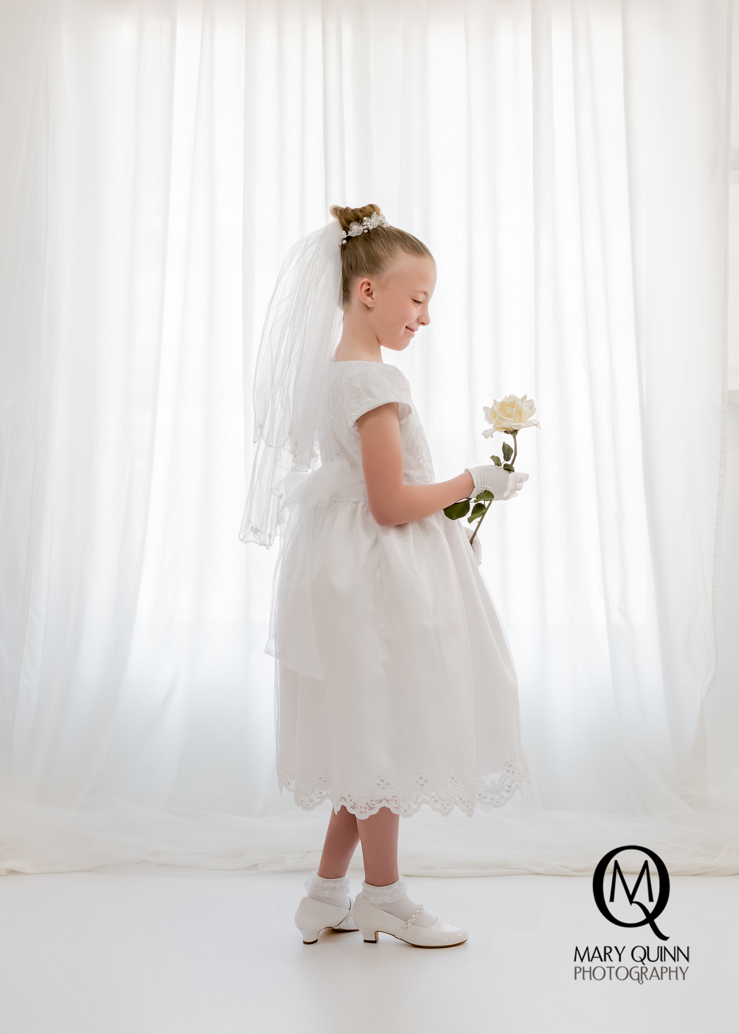 Image of a Communion girl standing in front of white curtains photographed by Mary Quinn.